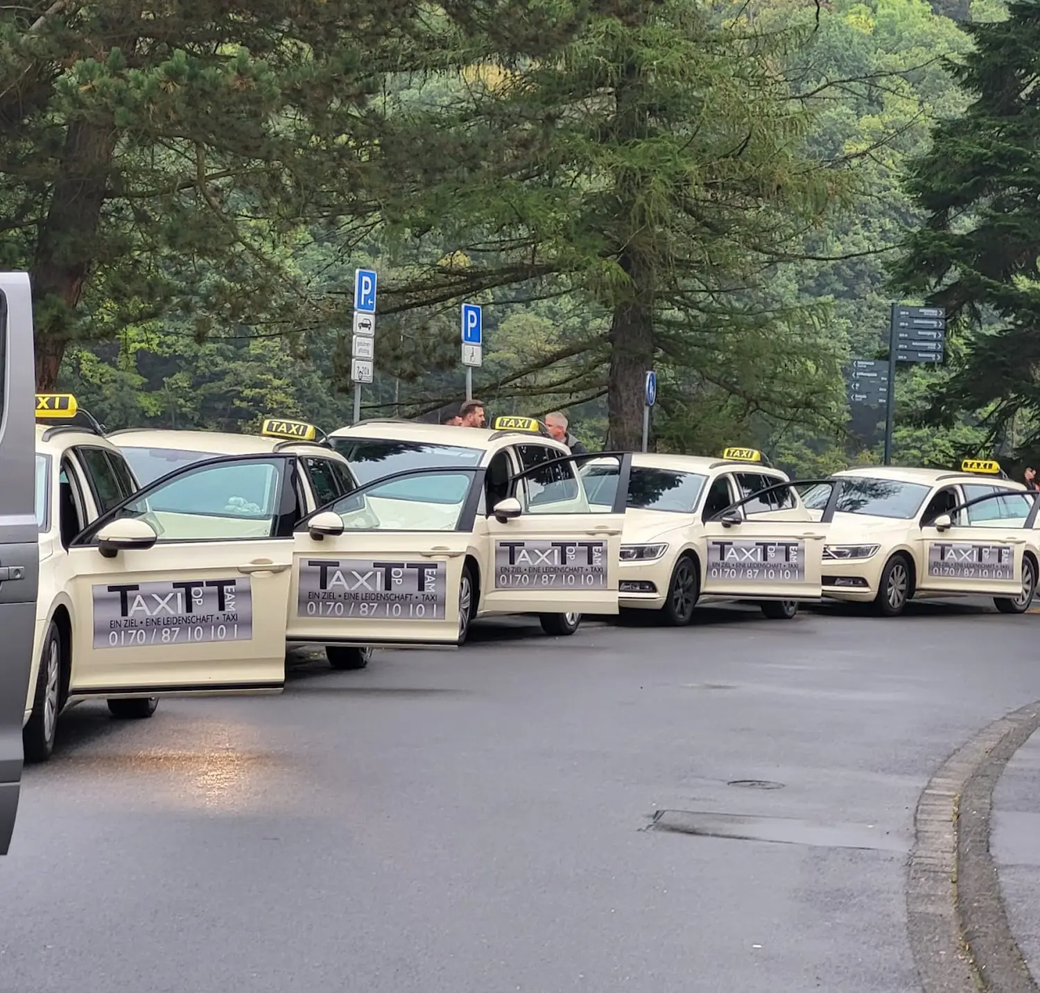 BonChance & TaxiTopTeam GmbH - Transportdienste - Eine Gruppe von fünf Personen, vermutlich Angestellte des Taxiunternehmens "AXI TT", posieren im Freien neben einem weißen Taxiwagen mit Firmenlogo für ein Foto.
