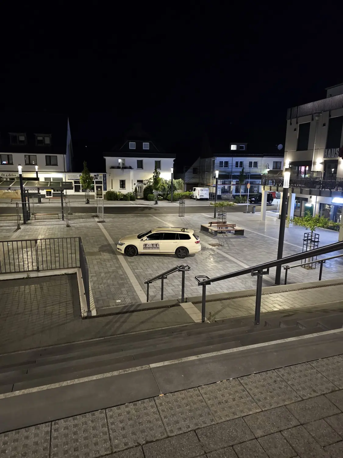 BonChance & TaxiTopTeam GmbH - Transportdienste - Ein weißes Taxi parkt nachts auf einem Stadtplatz oder einer Straße; im Hintergrund sind Gebäude und Treppen zu sehen.